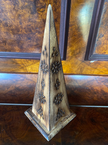 A decorative wooden pyramid-shaped object with carved floral or starburst motifs, standing on a matching square base against a polished wood-paneled background.