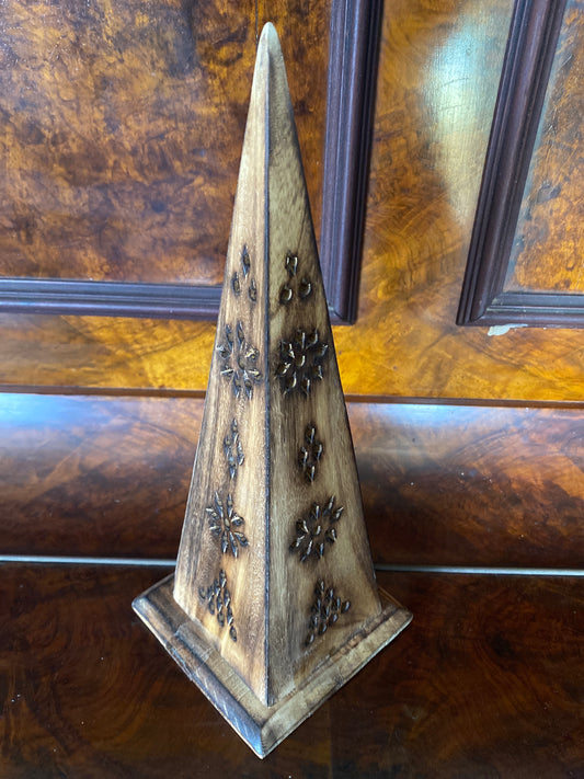 A decorative wooden pyramid-shaped object with carved floral or starburst motifs, standing on a matching square base against a polished wood-paneled background.