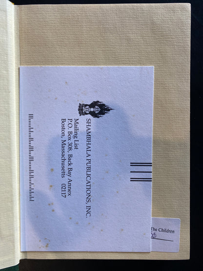 A vintage paper bookmark or insert from Shambala Publications, Inc., dated 1997, featuring a black-and-white illustration of a fairy and printed address details, placed on a beige textured book cover.