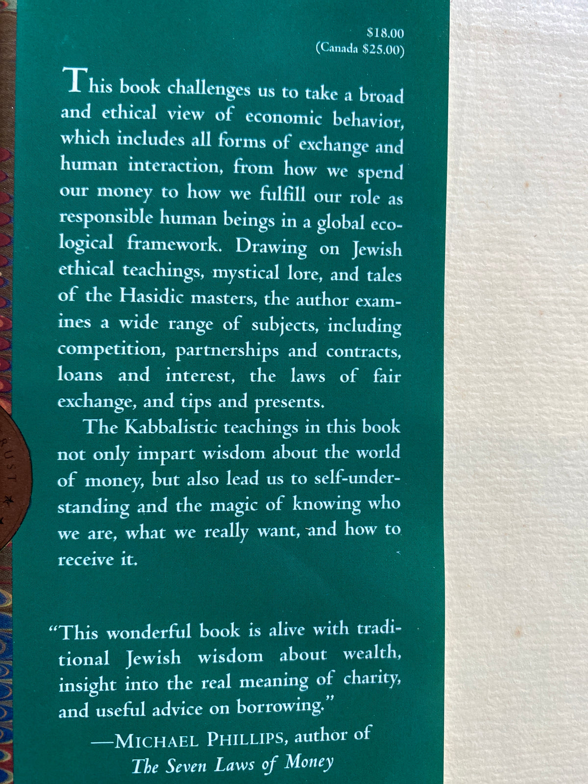 A book cover or printed page with a teal-green background and white text, photographed against a light beige textured surface. A partial decorative border with blue, gold, and red details is visible along the left edge.