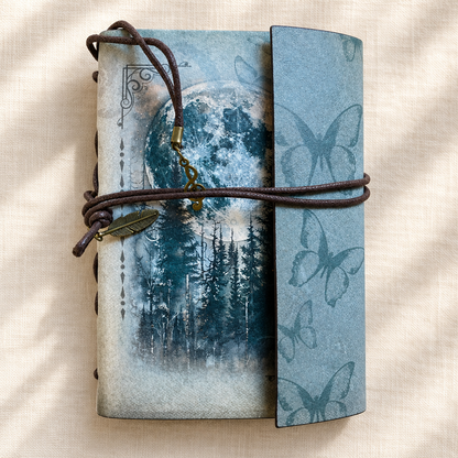 Blue-grey moonlit forest butterfly wrap journal with feather charm on a warm linen background; pen included but not shown